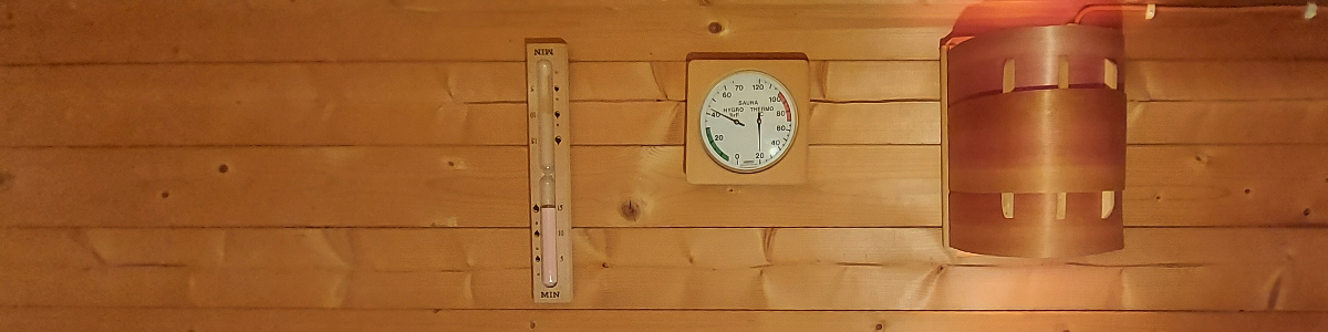 Unser Saunaofen Leistung Bild zeigt die Wand einer Saunakabine. An ihr hängen Beleuchtung, eine Sanduhr und ein Thermo-Hygrometer.